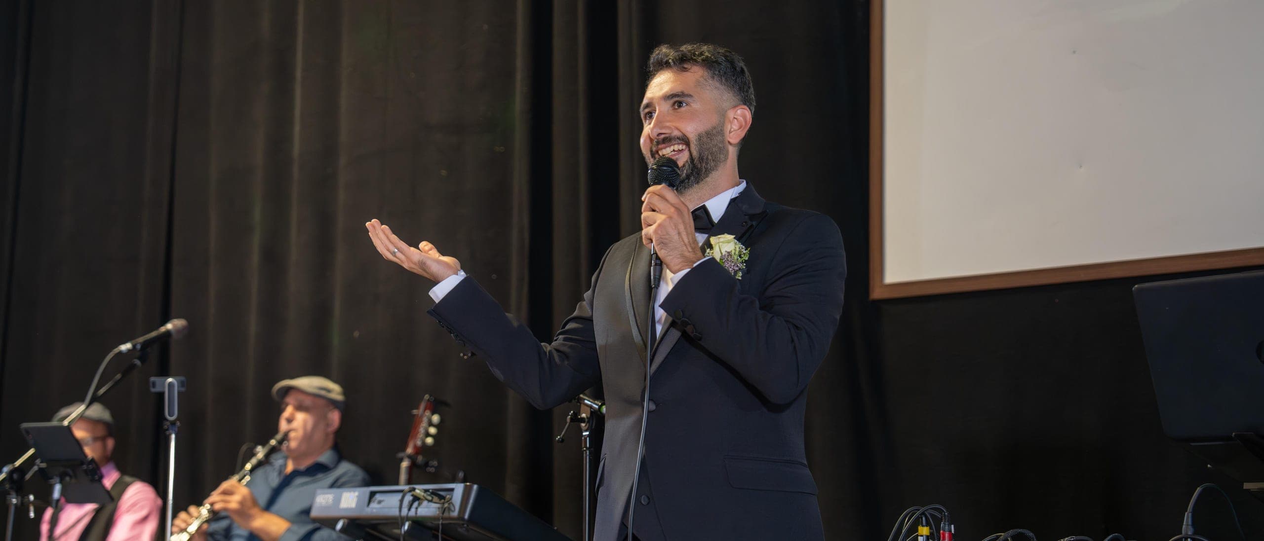 Veysel Durmuş moderiert mit Mikrofon auf der Bühne, Live-Musiker im Hintergrund