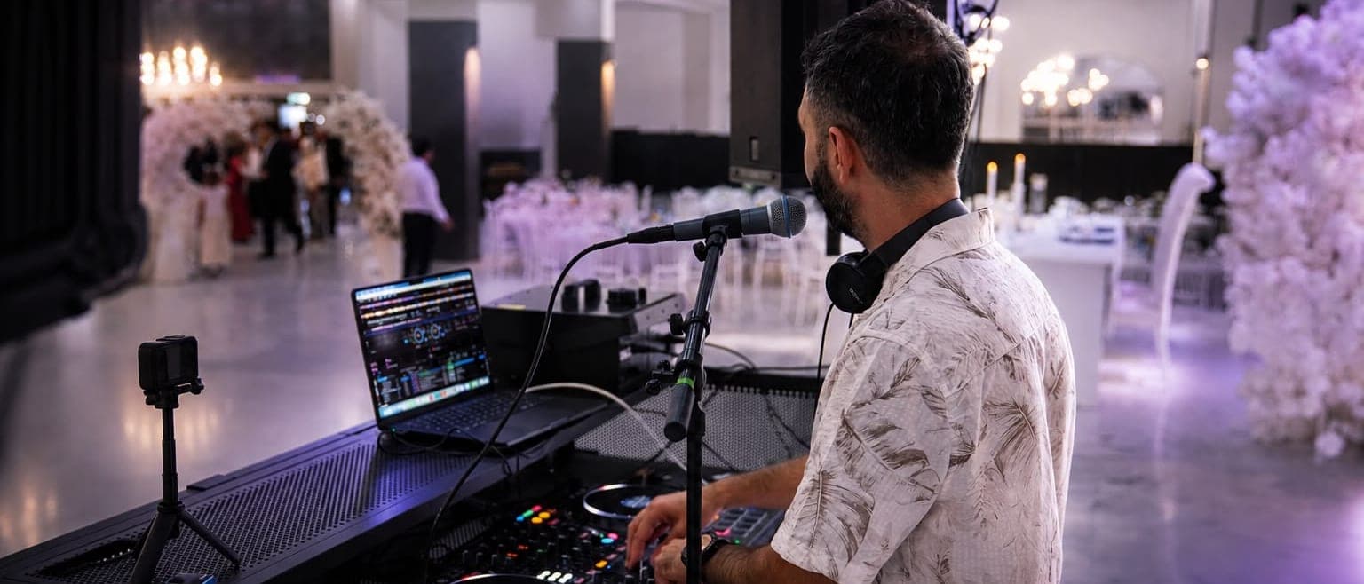 Hochzeitssaal am Abend: eingedeckte Tafeln und Blumenbogen im violetten Licht, Veysel Durmuş am DJ-Pult im Vordergrund