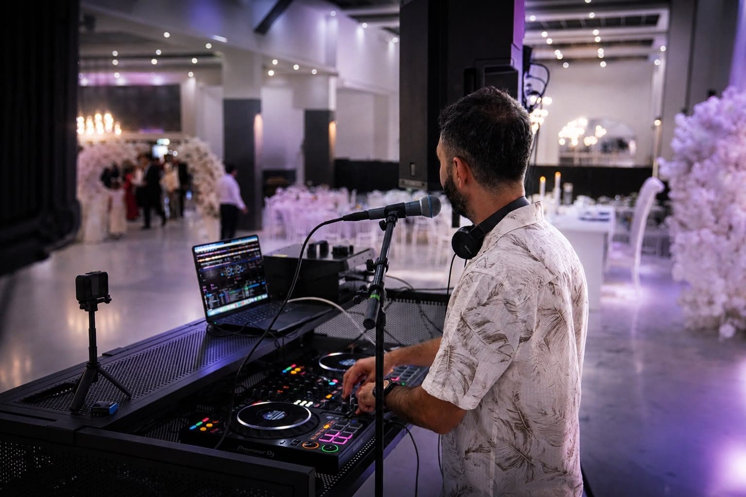 Hochzeitssaal am Abend: eingedeckte Tafeln und Blumenbogen im violetten Licht, Veysel Durmuş am DJ-Pult im Vordergrund