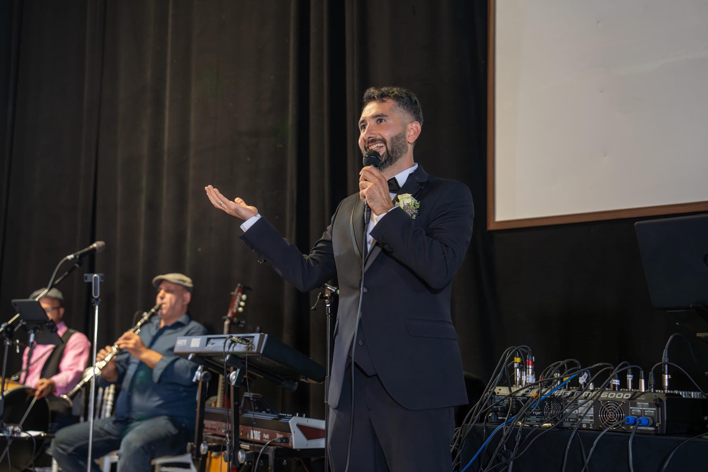 Veysel Durmuş moderiert mit Mikrofon, Live-Musiker mit Klarinette im Hintergrund
