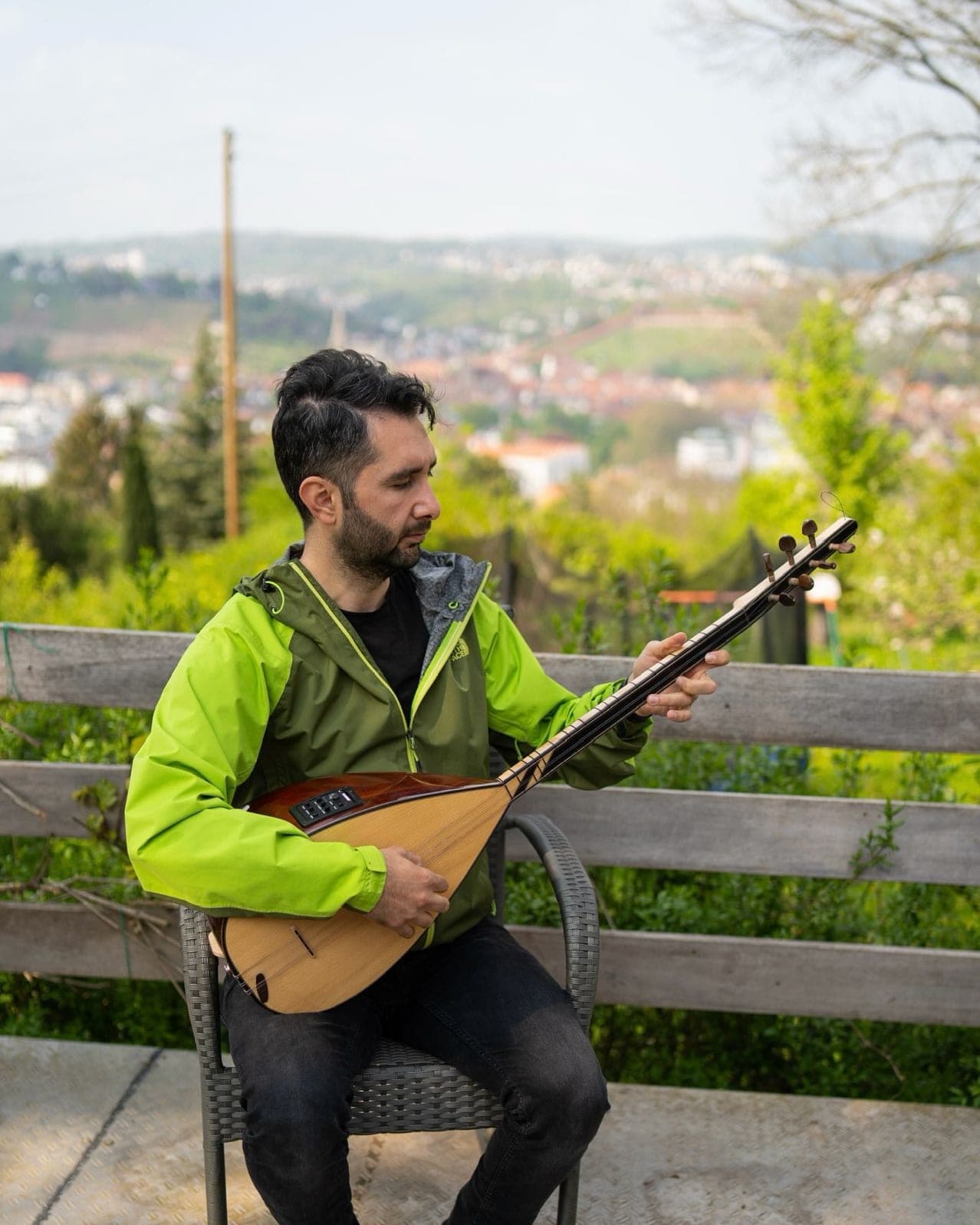 Veysel Durmuş spielt Bağlama auf einer Bank im Freien, Weinberge und Ortschaft im Hintergrund